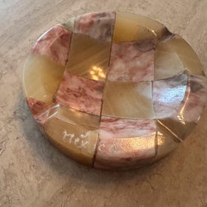 May include: A round, mosaic-style ashtray with a checkerboard pattern. The ashtray is made of polished stone in shades of beige, pink, and brown. The word "Mex" is etched on the side. A decorative item.