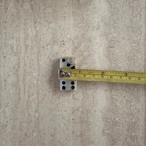May include: A pair of silver dominoes with black dots is measured with a yellow tape measure. The dominoes are approximately 1 inch in length. The tape measure shows the measurement in inches.