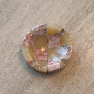 May include: A round, decorative stone coaster with a checkerboard pattern. The squares alternate between light yellow and pinkish-red hues. The word "Mex" is etched on the bottom right corner.