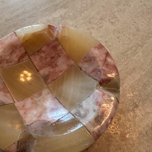 May include: A round, mosaic-style dish with a patchwork design. The dish features a combination of beige, cream, and pink-toned squares. The surface has a glossy finish, reflecting light.