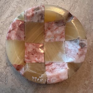 May include: A round, handcrafted stone coaster with a checkerboard pattern. The coaster is made of alternating squares of beige, pink, and cream-colored stone. The word "Mexico" is etched on the side.