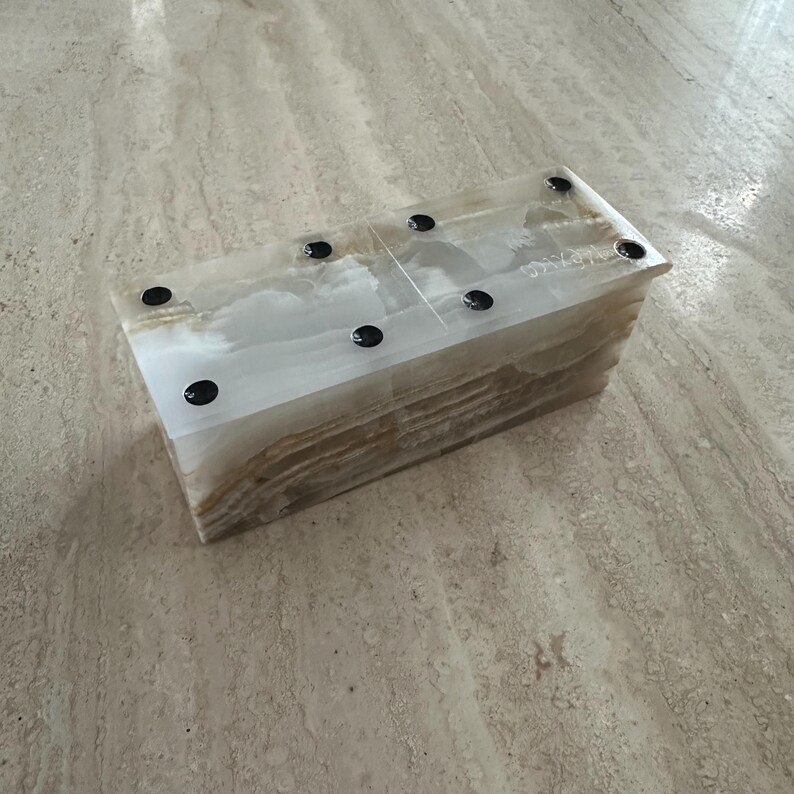 May include: Rectangular onyx box with a layered, translucent appearance. The box features a mix of white, beige, and brown tones, with six black circular accents on the top surface. The natural stone has a polished finish.