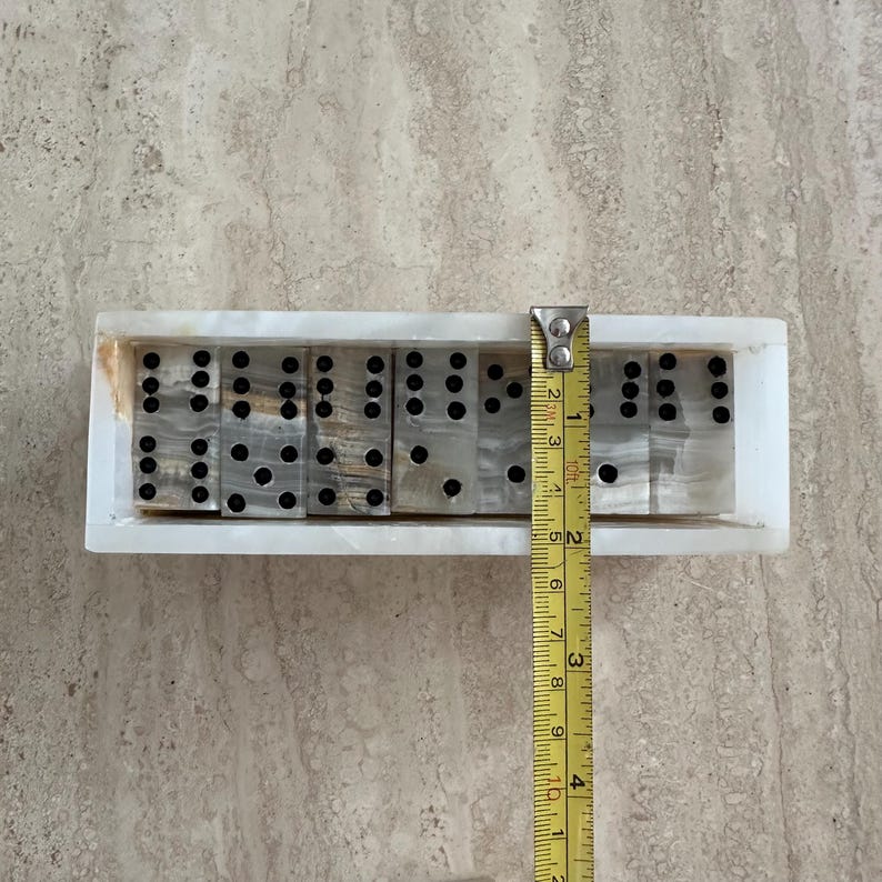 May include: A set of dominoes with black dots, resting in a rectangular white onyx holder. The dominoes are approximately 1 inch wide. A yellow measuring tape is next to the dominoes.