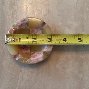May include: A round, mosaic-style coaster with a checkerboard pattern of beige, pink, and tan stone pieces. The coaster is approximately 4 inches in diameter. The word "Mexico" is etched on the side.