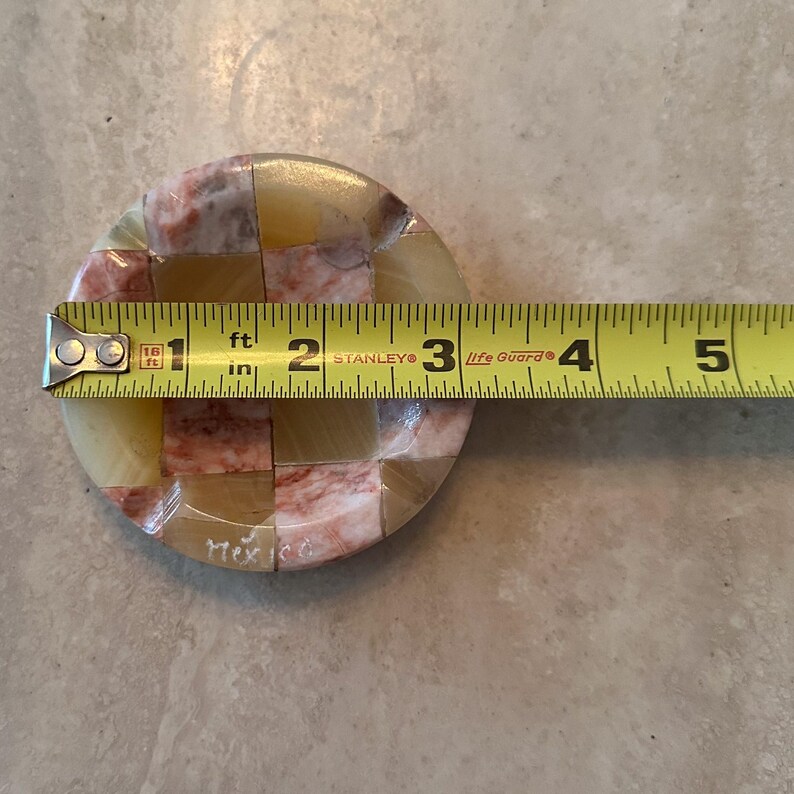 May include: A round, mosaic-style coaster with a checkerboard pattern of beige, pink, and tan stone pieces. The coaster is approximately 4 inches in diameter. The word "Mexico" is etched on the side.