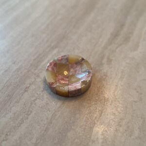 May include: A round, decorative ashtray with a checkerboard pattern of tan, pink, and cream-colored stone. The ashtray has a glossy finish and is set against a neutral background.