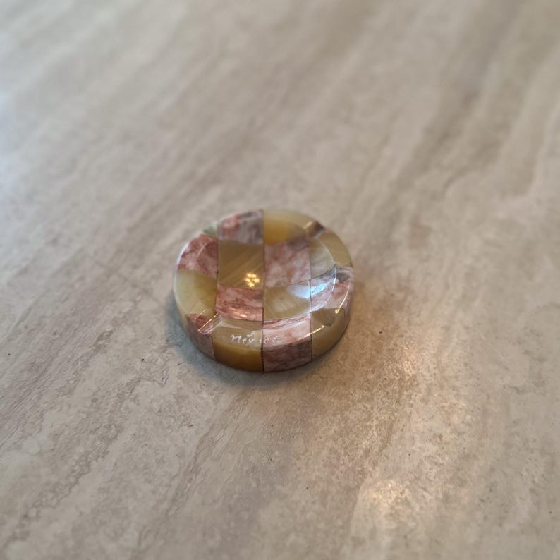 May include: A round, decorative ashtray with a checkerboard pattern of tan, pink, and cream-colored stone. The ashtray has a glossy finish and is set against a neutral background.