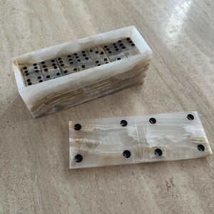 May include: A set of dominoes in a rectangular, layered onyx box. The dominoes are black and white, with black dots indicating their values. The box is open, revealing the dominoes inside, and a single domino is placed outside the box.