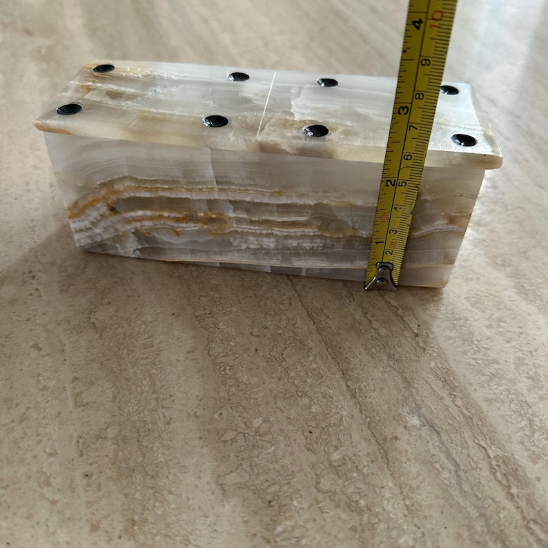 May include: A rectangular onyx box with a layered, translucent appearance. The box is approximately 4 inches tall. The top has six small, black, circular accents. The box is on a light-colored surface.
