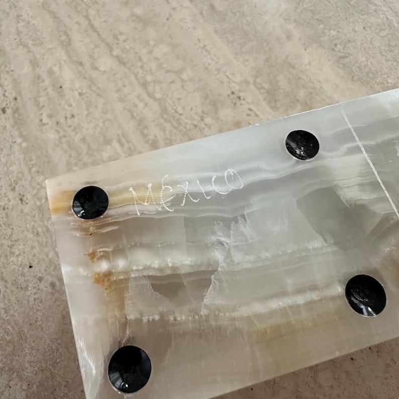 May include: A rectangular, translucent onyx piece with the word "Mexico" etched on the surface. The onyx has a layered, natural pattern with shades of white, cream, and tan. Four black, circular feet are attached to the bottom.