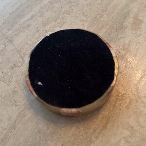 May include: A round, decorative object with a black, velvety top and a light-colored, patterned rim. The rim features shades of beige, pink, and gold. The object is set against a neutral, textured background.