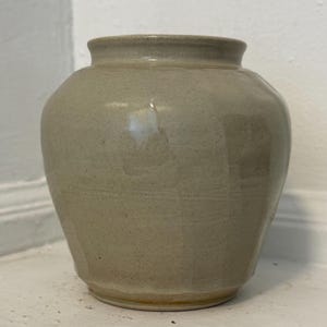 May include: A small, round ceramic vase with a smooth, glossy, light beige glaze. The vessel features a wide, bulbous body that tapers toward a narrow, circular opening at the top, showcasing a subtle, speckled texture within the clay finish.