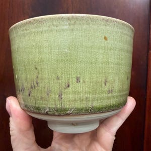 May include: A ceramic bowl with a light green crackle glaze finish. The exterior features a subtle vertical texture and dark brown speckling near the base, which transitions into a smooth, unglazed white footed bottom.
