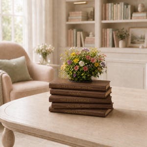 May include: A stack of five brown books with gold lettering titled The Old Man and the Sea, Walden, The Little Prince, Into the Wild, and Sophia's Library, topped with a vibrant arrangement of small yellow, pink, white, and purple wildflowers.