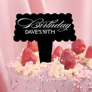 May include: A black scalloped cake topper featuring the white text "Birthday" in a script font and "DAVE'S 50TH" in a sans-serif font, inserted into a light pink frosted cake decorated with fresh red strawberries and small white pearl sprinkles.