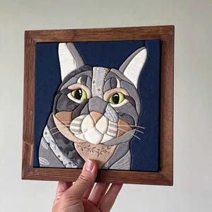 Könnte beinhalten: Ein quadratisches, gerahmtes Stoffkunstwerk mit einem stilisierten Katzenporträt. Die Katze besteht aus verschiedenen grauen, weißen und beigen Stoffstücken mit komplizierten Mustern vor einem einfarbigen marineblauen Hintergrund in einem dunklen Holzrahmen.