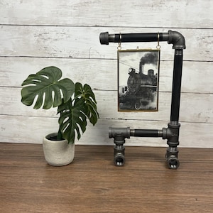 May include: A black and gray industrial pipe stand featuring a rectangular brass-framed picture of a steam locomotive. Beside the stand is a green monstera plant in a small, textured, light-colored ceramic pot.