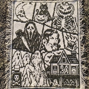 May include: A rectangular crochet blanket featuring a dark green and cream tapestry design of Halloween motifs including a skull, a cat, a pumpkin, a ghost-like figure, a haunted house, and a bottle with a skull and crossbones label.