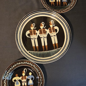 May include: Three round ceramic plates featuring a black background with a central illustration of three figures in white, brown, and blue attire. The plates have concentric circular borders in white, blue, and brown with decorative patterns.