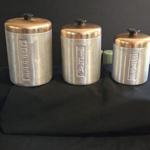 May include: Three cylindrical aluminum canisters with copper-toned lids and black knobs. The canisters feature embossed vertical text labels reading COFFEE, TEA, and GREASE, arranged in descending order of size from left to right.
