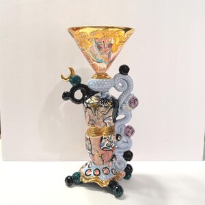 May include: A decorative ceramic chalice featuring a wide gold-rimmed bowl with painted faces, a stacked body with blue polka-dot patterns, and a sculptural side handle adorned with black, pink, and blue spheres and a crescent moon accent.