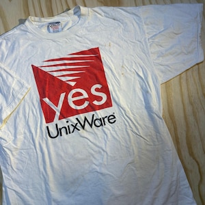 Puede incluir: Una camiseta blanca de manga corta con un gráfico de un cuadrado rojo con líneas blancas horizontales y la palabra yes en letras minúsculas blancas. Debajo del cuadrado rojo, el texto UnixWare está impreso en negro. La camiseta tiene una etiqueta Oneita.