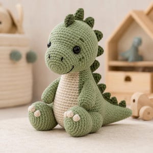 May include: A sage green and cream colored crochet dinosaur stuffed animal. The toy features a rounded head, black plastic eyes, a textured spine with small triangular spikes, and a light cream belly. It is sitting in an upright position.