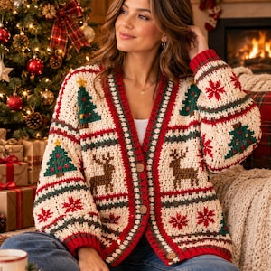 May include: A cream-coloured crochet cardigan featuring a festive holiday pattern with green Christmas trees, brown reindeer, and red snowflakes. The garment has a V-neckline, long sleeves, a red ribbed trim, and three round wooden buttons down the front.