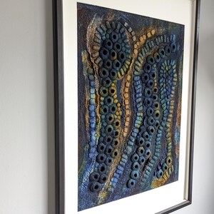 May include: A framed textile wall art piece featuring a textured, three-dimensional surface with organic, circular, and tubular shapes in shades of deep blue, gold, and dark brown, set within a simple black frame and a wide off-white mat board.