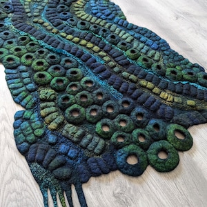 Puede incluir: Un tapiz de pared hecho de lana fieltrada en tonos azul profundo, verde azulado y verde bosque. El textil presenta una textura compleja y elevada con numerosas formas circulares tipo anillo y patrones lineales segmentados, suspendido de una varilla de madera.