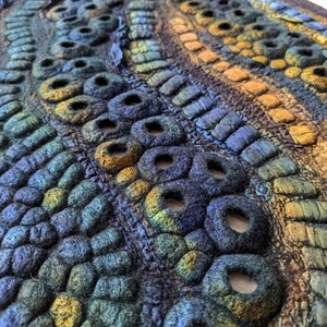 May include: A textile wall art piece featuring a dense, three-dimensional surface of felted wool. The composition consists of undulating rows of circular, hollow, and tubular shapes in a gradient of deep blue, teal, green, and golden yellow tones.