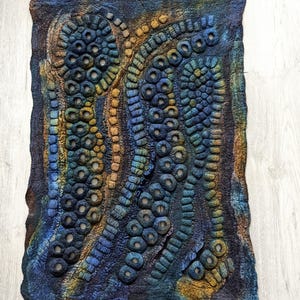 May include: A rectangular felted wall hanging featuring a textured, abstract design of raised circular and tubular shapes. The piece displays a gradient of deep blue, teal, and earthy ochre tones, with open circular voids integrated into the felted surface.
