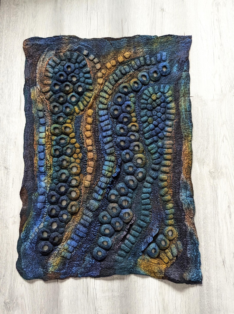 May include: A rectangular felted wall hanging featuring a textured, abstract design of raised circular and tubular shapes. The piece displays a gradient of deep blue, teal, and earthy ochre tones, with open circular voids integrated into the felted surface.