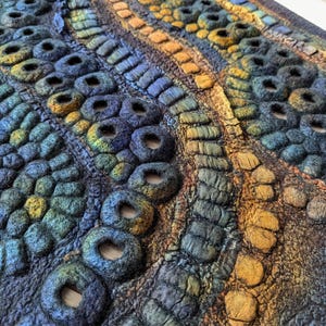 May include: A textured fiber art piece featuring a complex arrangement of felted circular forms and raised linear patterns. The surface displays a gradient of deep blues, forest greens, and warm ochre tones, creating a dense, tactile, and organic relief.