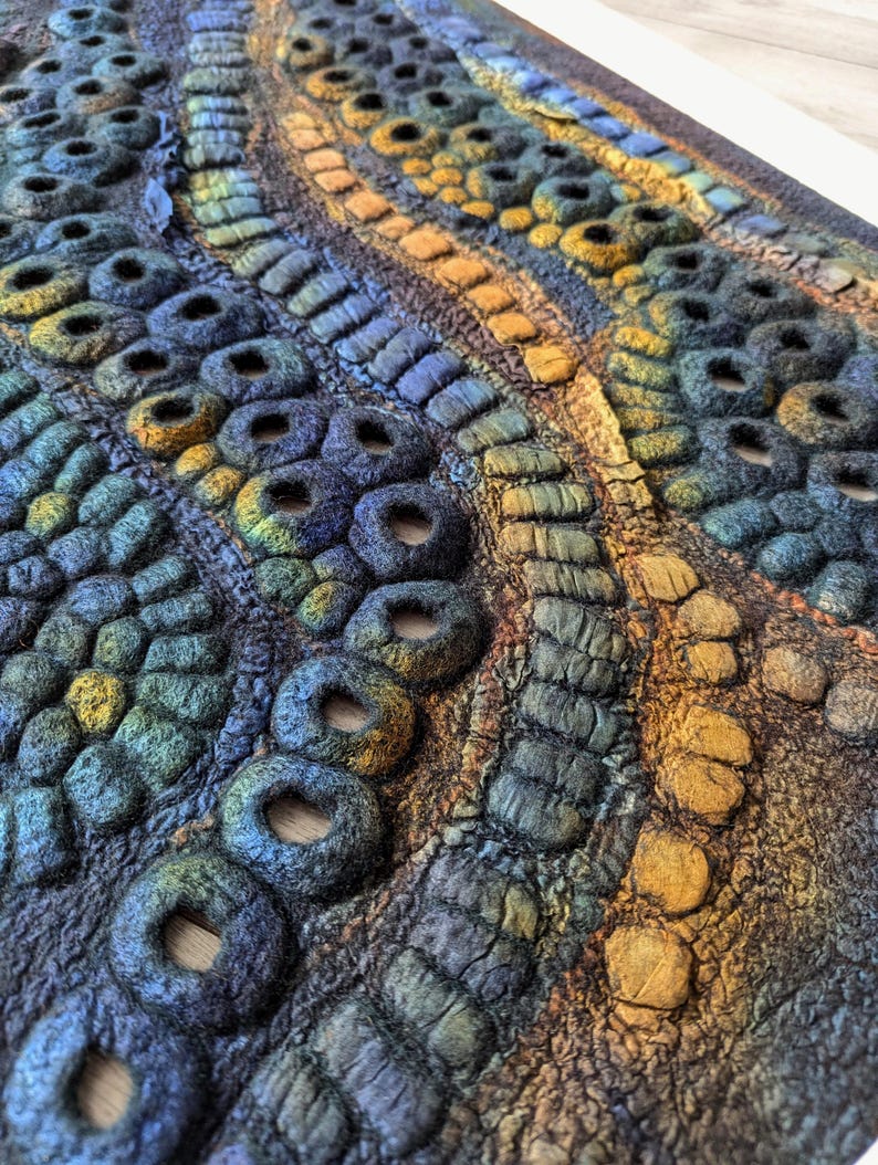 May include: A textured fiber art piece featuring a complex arrangement of felted circular forms and raised linear patterns. The surface displays a gradient of deep blues, forest greens, and warm ochre tones, creating a dense, tactile, and organic relief.