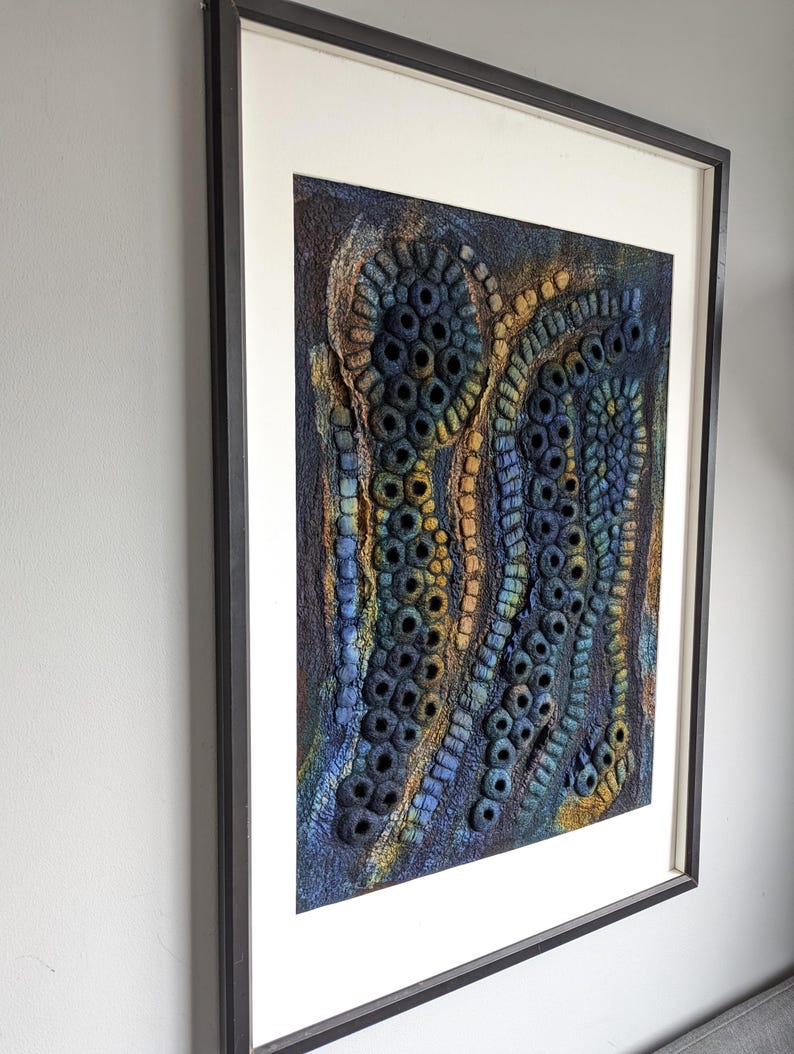 May include: A framed textile wall art piece featuring a textured, three-dimensional surface with organic, circular, and tubular shapes in shades of deep blue, gold, and dark brown, set within a simple black frame and a wide off-white mat board.