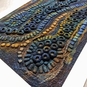 May include: A textured wall art piece featuring a raised, organic pattern of circular and tubular shapes in shades of deep blue, teal, and golden yellow. The composition consists of dense, repeating clusters and wavy lines set within a white mat.