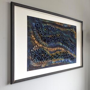 May include: A rectangular framed wall art piece featuring a textured, three-dimensional fiber art composition. The artwork displays undulating, wave-like patterns in deep shades of blue, navy, and gold, created with numerous small, circular, and tubular felted elements.