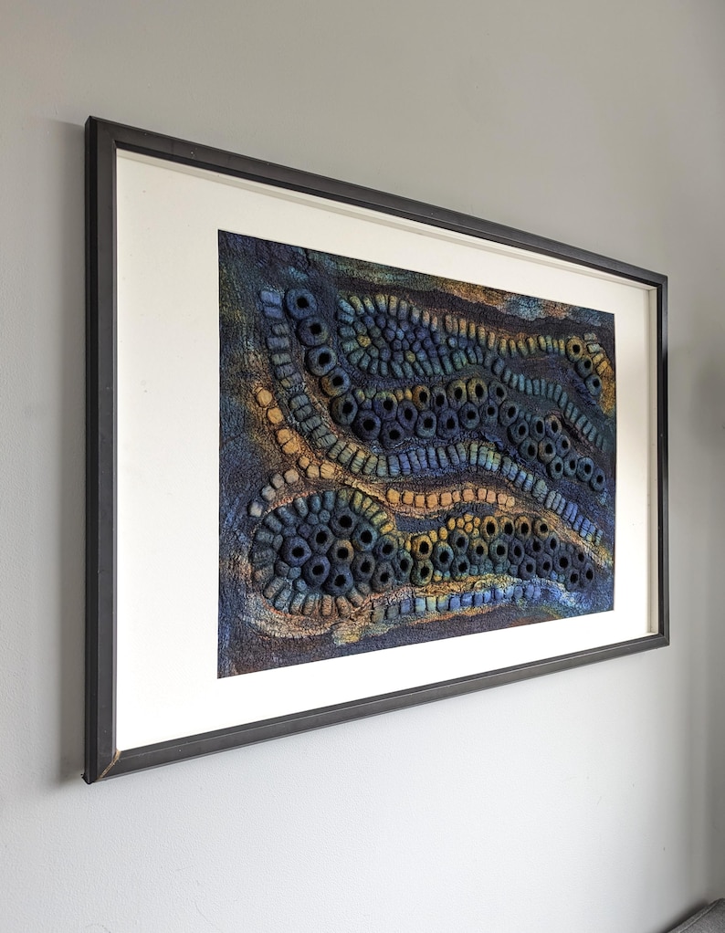 May include: A rectangular framed wall art piece featuring a textured, three-dimensional fiber art composition. The artwork displays undulating, wave-like patterns in deep shades of blue, navy, and gold, created with numerous small, circular, and tubular felted elements.