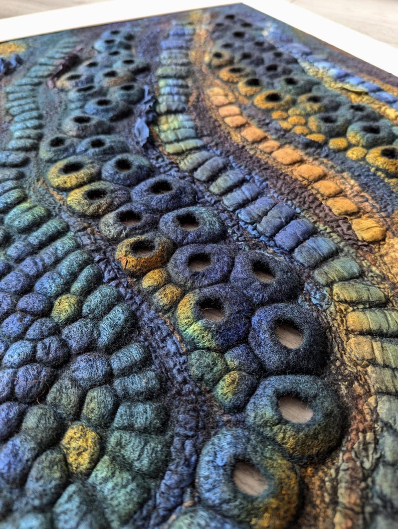 May include: A textile wall art piece featuring a dense, three-dimensional surface of felted wool. The composition consists of undulating rows of circular, hollow, and tubular shapes in a gradient of deep blue, teal, green, and golden yellow tones.