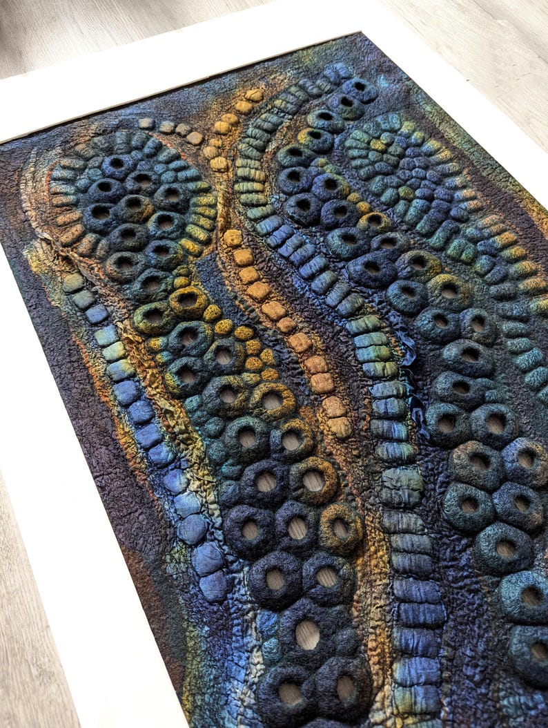 May include: A rectangular fibre art wall hanging featuring a textured, three-dimensional surface with raised circular and linear patterns. The piece displays a gradient of deep blue, teal, and golden yellow tones, framed within a white border.