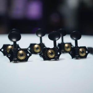 Pode incluir: Um conjunto de tarraxas de guitarra pretas com engrenagens expostas na cor latão e botões ovais pretos. Cada tarraxa possui uma placa de montagem com dois orifícios para parafusos e um mecanismo central de engrenagem para ajustar as cordas.