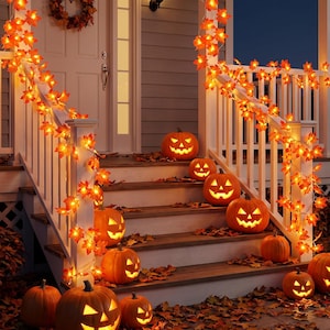Peut inclure: Guirlandes de feuilles d'érable artificielles illuminées par des lumières LED orange chaud, drapées sur des rampes de porche blanches, accompagnées de plusieurs citrouilles sculptées en jack-o'-lantern aux visages lumineux sur les marches.