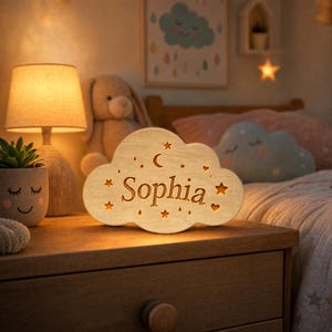 Pode incluir: Uma luz noturna de madeira em forma de nuvem com o nome Sophia gravado no centro. A luz é iluminada por dentro, emitindo um brilho quente através de recortes em forma de estrelas, corações e uma lua crescente ao redor do nome.