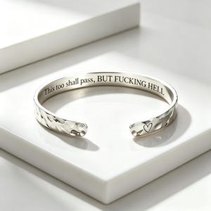 May include: A silver-toned cuff bracelet with a hammered exterior texture. The interior is engraved with the text "This too shall pass, BUT FUCKING HELL" in a serif font. One end of the bracelet features a small, simple engraved heart symbol.