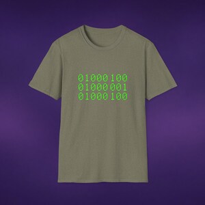 May include: A sage green short-sleeved t-shirt featuring a graphic print of three rows of binary code in a bright lime green colour. The text reads 01000100, 01000001, and 01000100, displayed in a pixelated font centred on the chest of the garment.