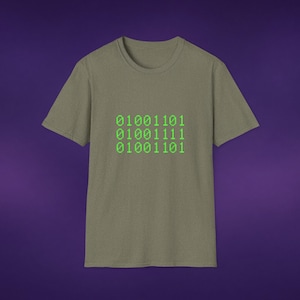 May include: An olive green short-sleeved t-shirt featuring a graphic print of three rows of bright green binary code. The text consists of the numbers 01001101, 01001111, and 01001101, displayed in a pixelated font centered on the chest of the garment.