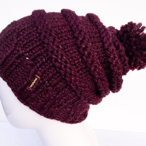 May include: A burgundy knitted beanie with a pom pom on top. The beanie has a small gold tag that says "The Hat Store".