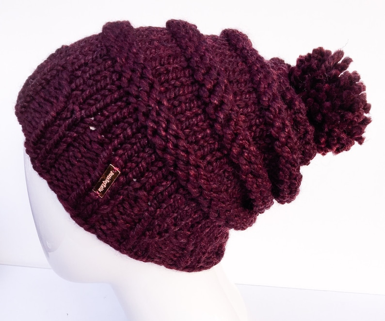 May include: A burgundy knitted beanie with a pom pom on top. The beanie has a small gold tag that says "The Hat Store".