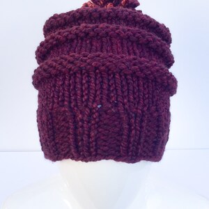 May include: A burgundy knitted beanie with a pom pom on top. The beanie has a cable knit pattern.